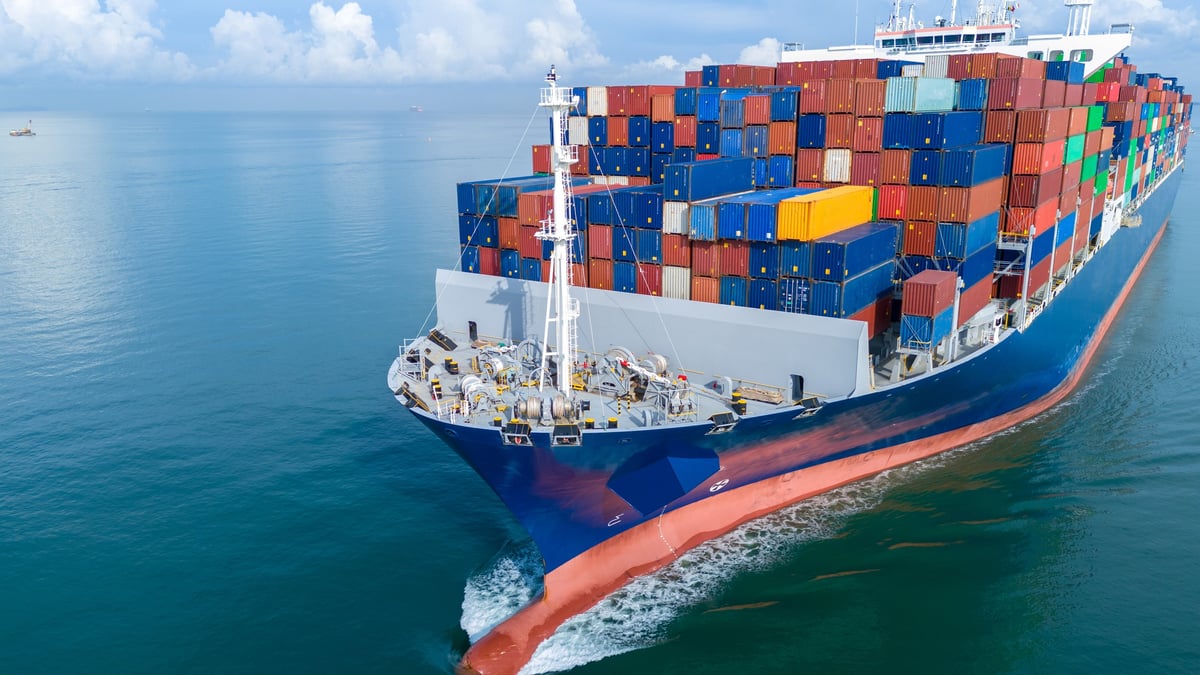 Aerial front view of a large cargo ship fully loaded with colorful shipping containers navigating through calm ocean water.
